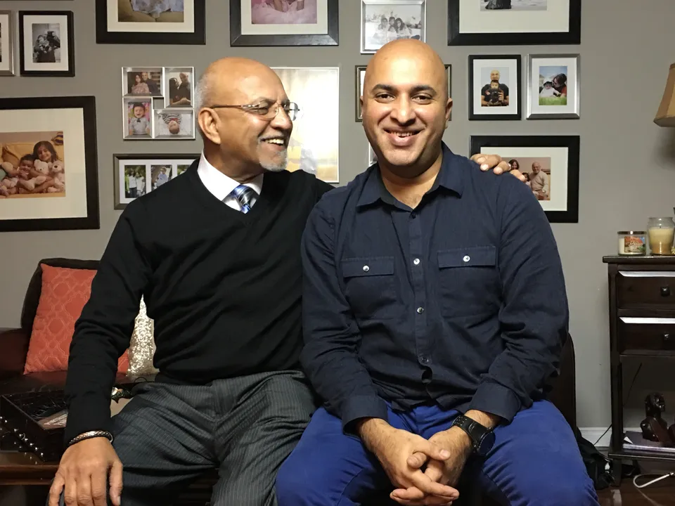 Rashid Mughal sits beside his son Imran in front of a wall of family photographs, an arm around his shoulders, laughing as Imran smiles