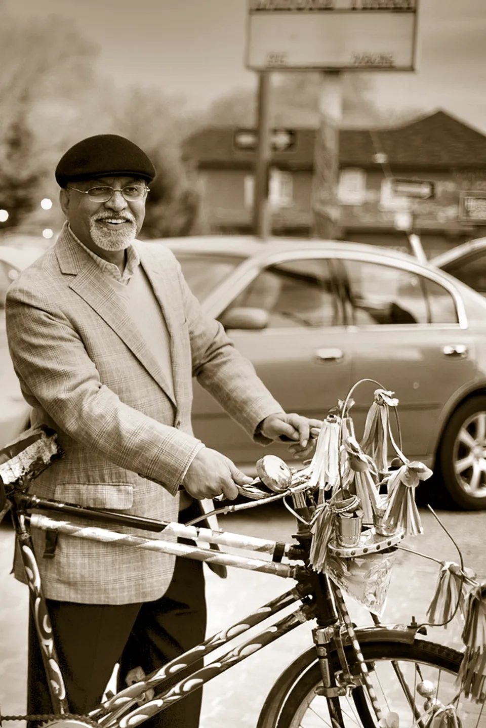 Rashid Mughal in a flat cap and glasses, smiling broadly on a city street beside a bicycle decorated with ribbons and tassels