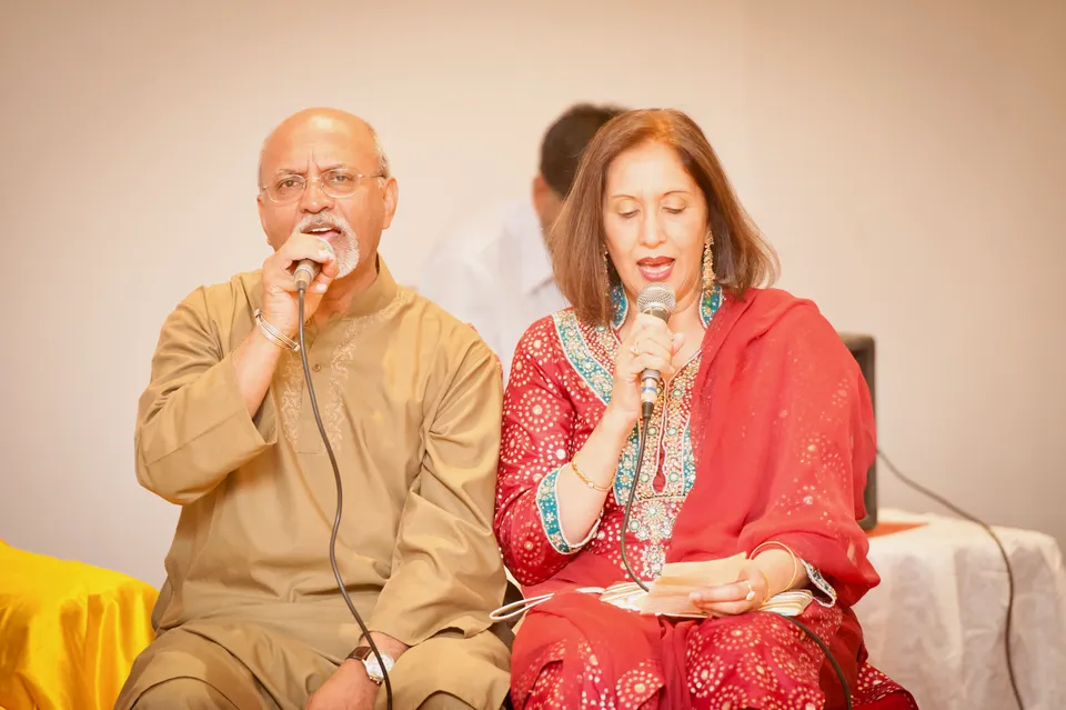 Rashid Mughal sings into a microphone at a gathering, his wife singing beside him in red, reading the verses from a slip of paper